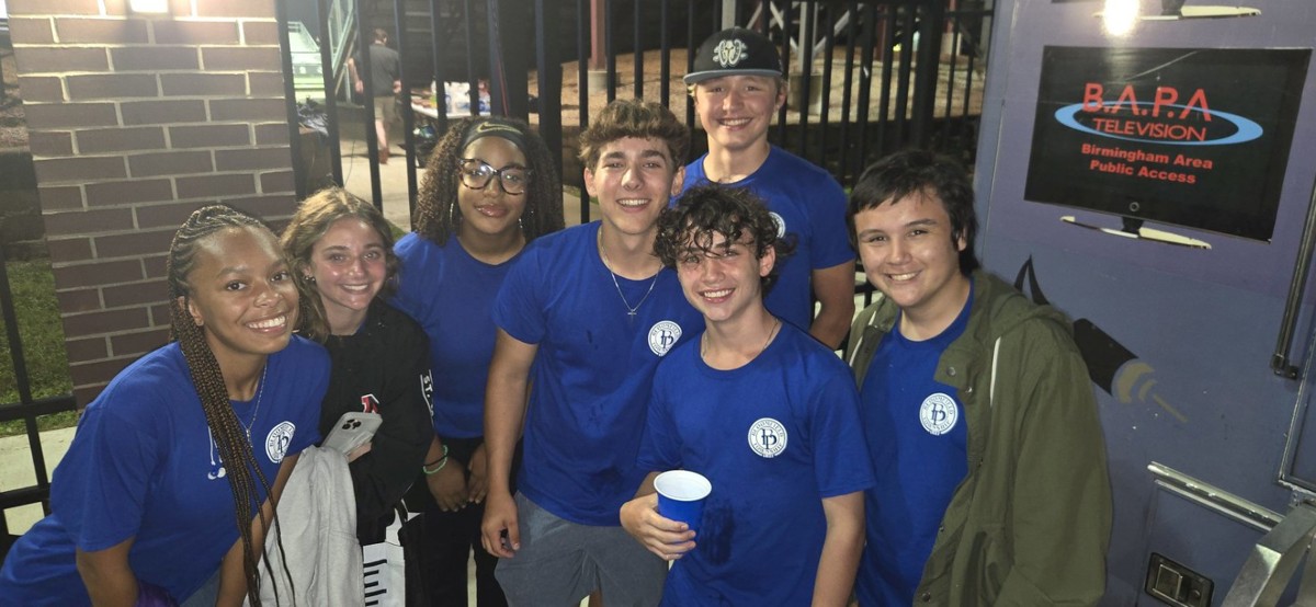 Group of 7 teens wearing Bloomfield Township logo t-shirts standing outside by a tv production truck in the evening.