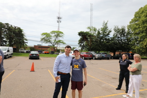 A man with his arm around a teenage boy smiling for camera in a parking lot with onlookers